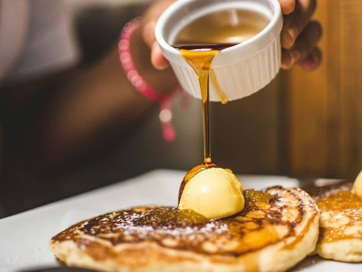 A person pours syrup on pancakes topped with butter, focusing on the syrup's smooth stream and the pancakes' texture.