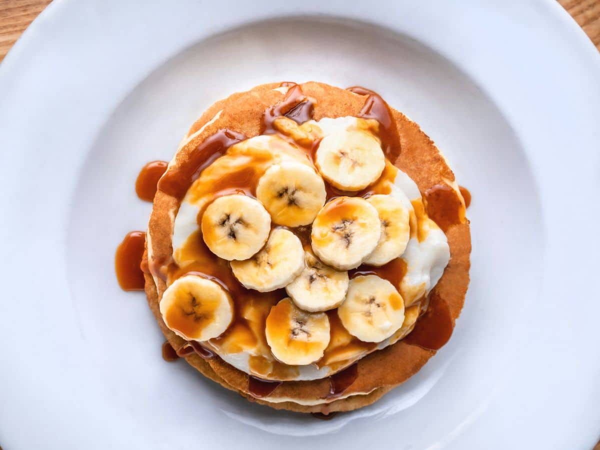 A stack of pancakes topped with sliced bananas and drizzled with caramel sauce, served on a white plate.