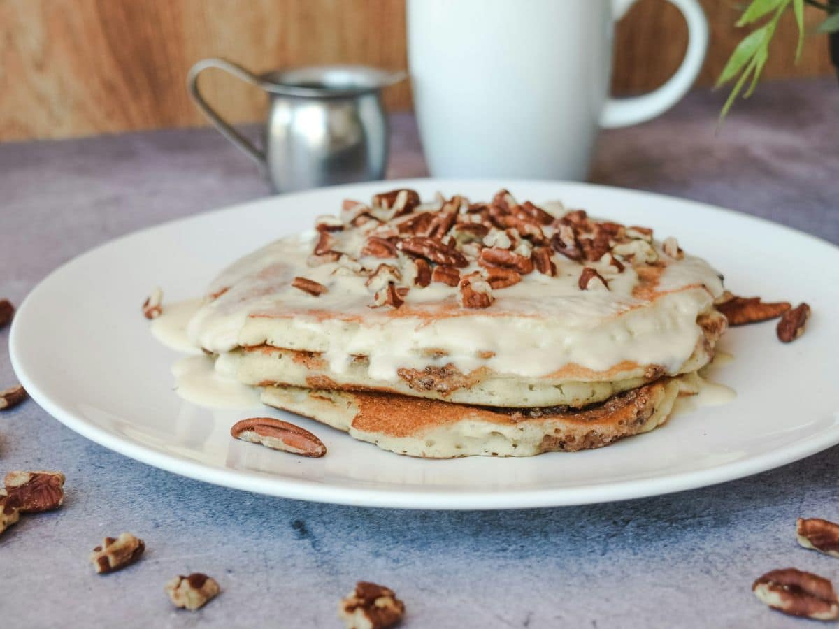 A stack of pancakes topped with cream and pecans on a white plate, with a milk jug in the background.