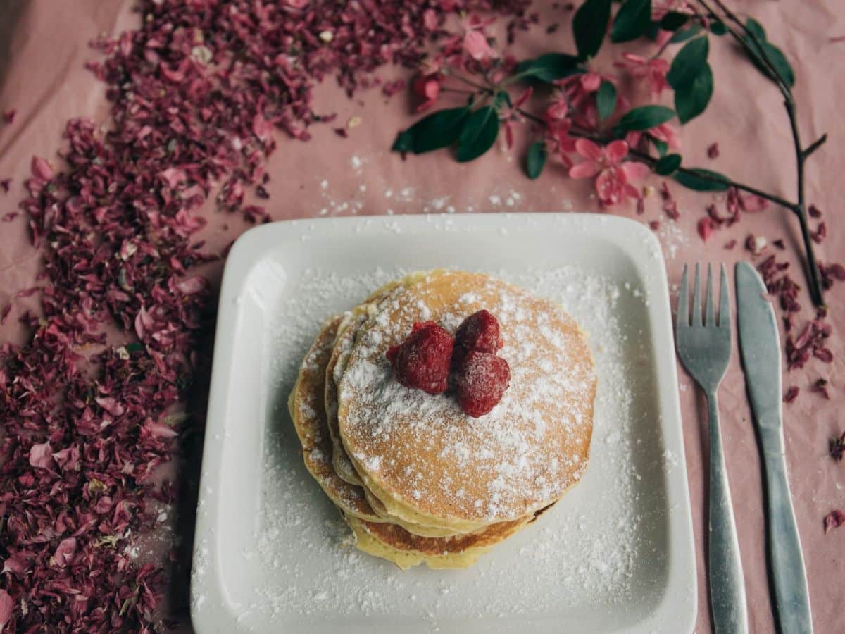 A stack of pancakes topped with raspberries and powdered sugar, served on a white plate surrounded by scattered pink petals, with a fork and knife on the side.