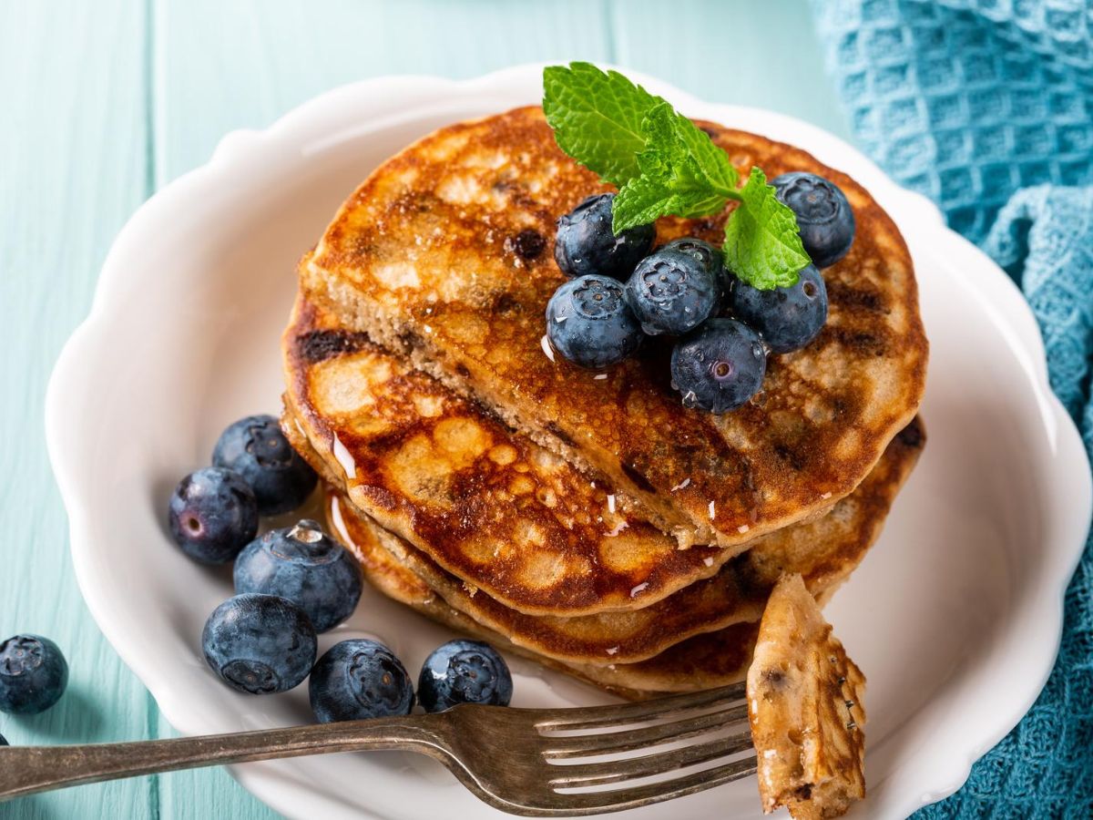 A stack of pancakes topped with fresh blueberries and a sprig of mint on a white plate, with a fork on the side.