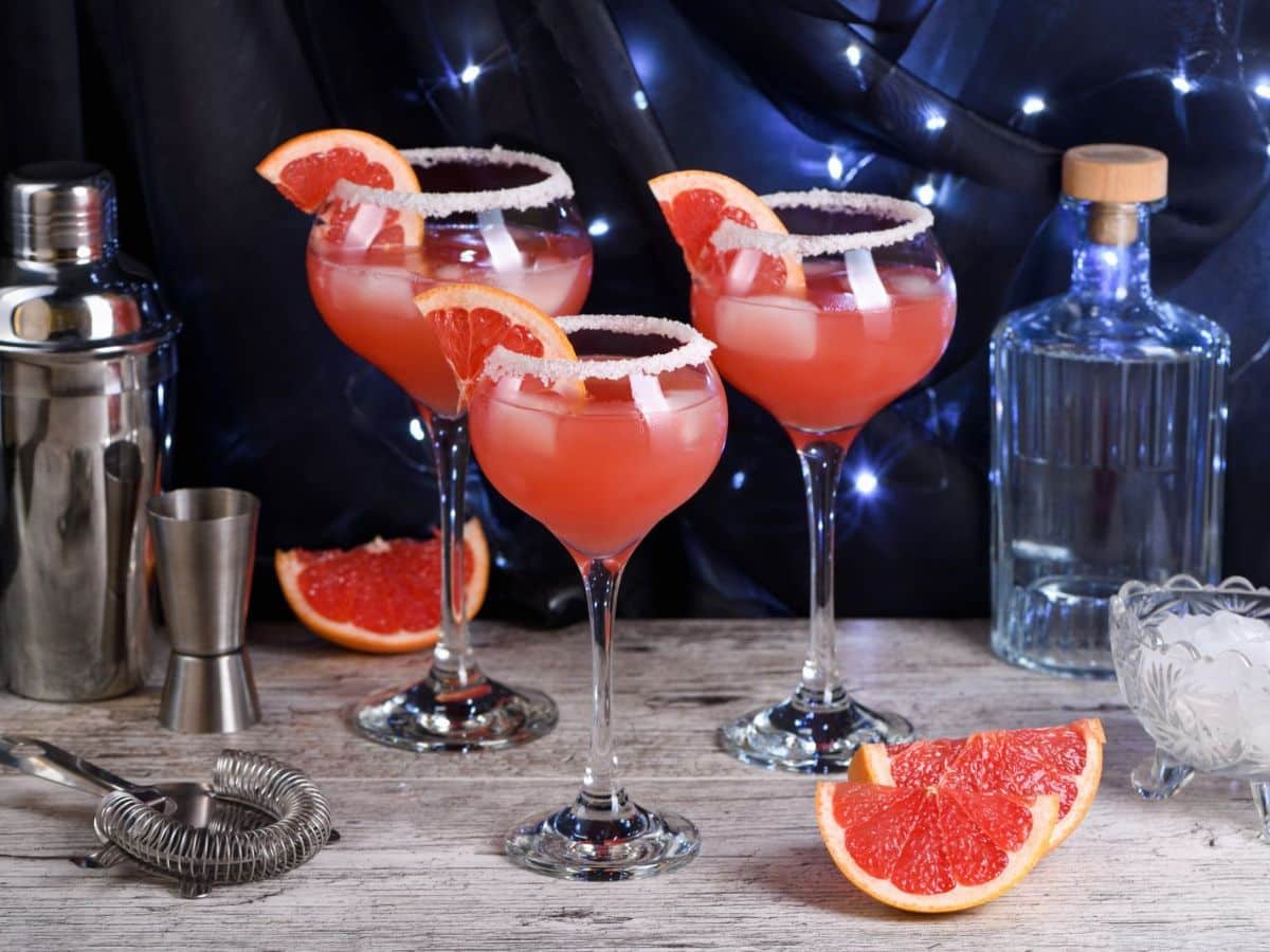 Three grapefruit cocktails with salt rims on a bar counter, surrounded by grapefruit slices, a cocktail shaker, and a bottle.