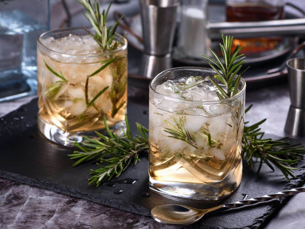 Two glasses of whiskey with ice and rosemary sprigs on a dark slate tray, bar tools in the background.