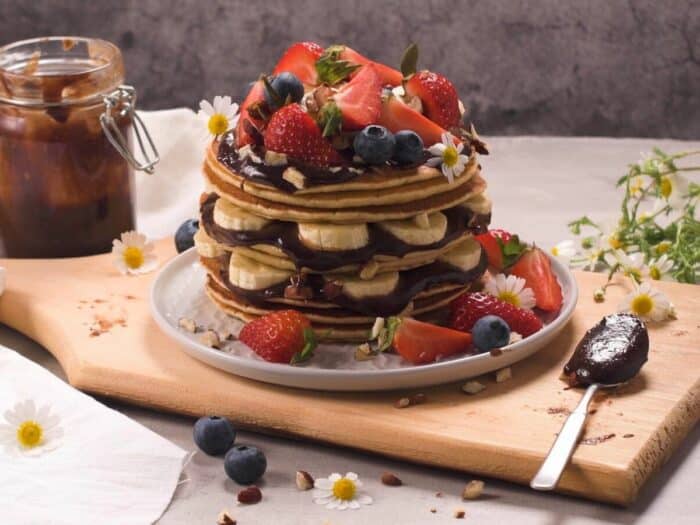 Stack of pancakes with strawberries, blueberries, and chocolate sauce on a wooden board, next to a jar of sauce and small flowers.